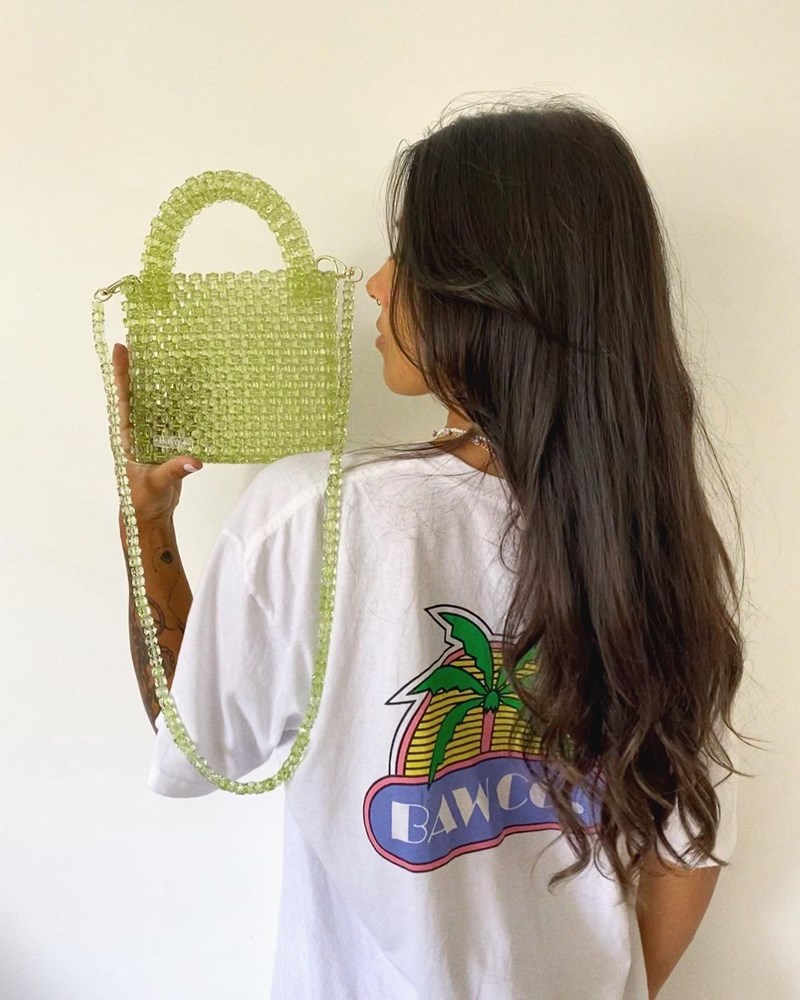 Modelo segura bolsa de miçangas verdes. Ela está de costas e veste uma camiseta branca com estampa - Metrópoles 