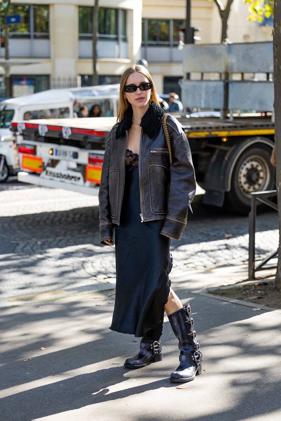 Mulher branca e jovem, de cabelo liso loiro, posa para foto na rua durante a Semana de Moda de Paris. Ela usa um vestido preto de cetim, com detalhes de renda; uma jaqueta de couro e uma bota preta, também de couro. - Metrópoles