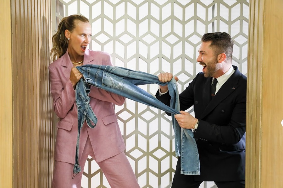 Os apresentadores Renata Kuerten e Lucas Anderi, do programa Esquadrão da Moda, posa para foto em cenário. Ela é uma mulher branca e jovem, de cabelo liso loiro, e usa um terno lilás. Ele é um homem branco e jovem, de cabelo e barba grislha, e usa terno completo cinza. - Metrópoles