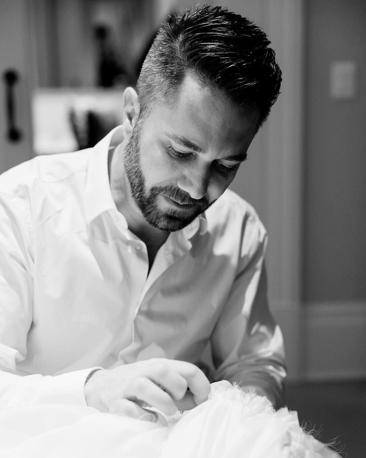 O estilista Lucas Anderi, apresentador do Esquadrão da Moda, do SBT, posa para foto enquanto costura. Ele é um homem branco e jovem, de cabelo e barba castanhos, e usa uma camisa branca de botão. - Metrópoles