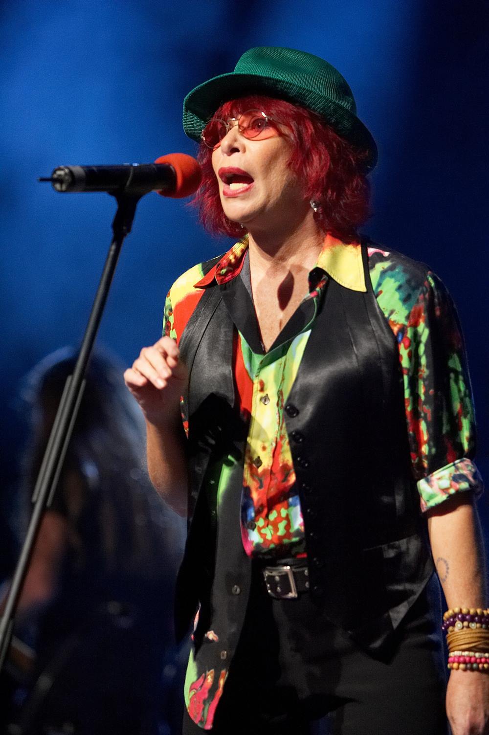 A cantora Rita Lee durante um show em 2008. Ela é uma mulher branca e idosa, com cabelos lisos pintados de vermelho. Usa um óculos amarelo, chapéu preto, uma camisa colorida e colete e calça, ambos pretos. - Metrópoles