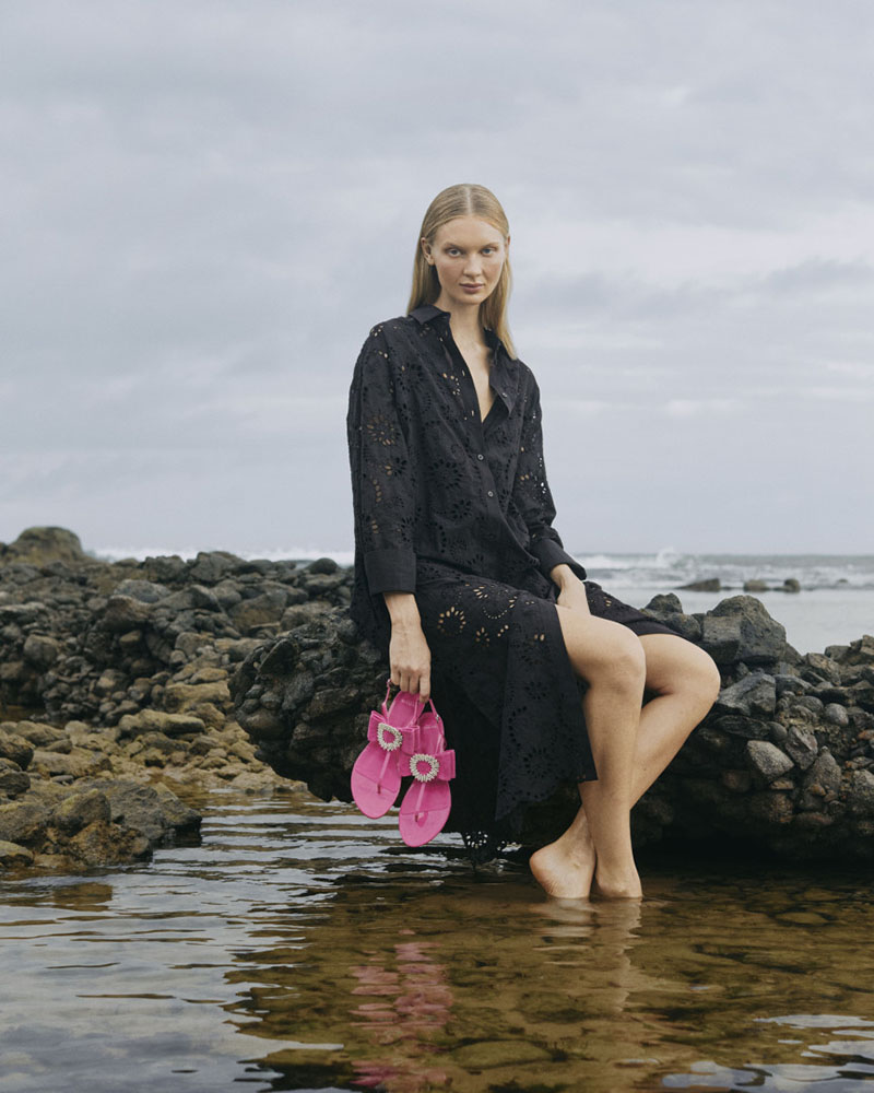 Foto de campanha publicitária da marca Alexandre Birman, do grupo Arezzo&Co. Na imagem uma modelo branca e jovem, de cabelos lisos loiros, posa para foto sentada na praia. Ela usa um vestido preto longo e segura, com as mãos, uma sandália rasteira rosa. - Metrópoles