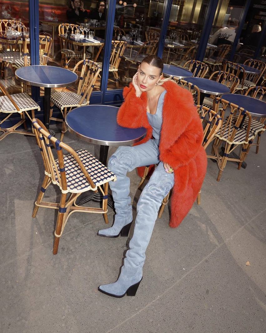 Foto de campanha da marca Paris Texas. Na imagem, uma mulher branca e jovem, de cabelo liso e castanho, usa um top, uma calça e uma bota, tudo com tecido jeans; e um casaco vermelho de pelo. - Metrópoles