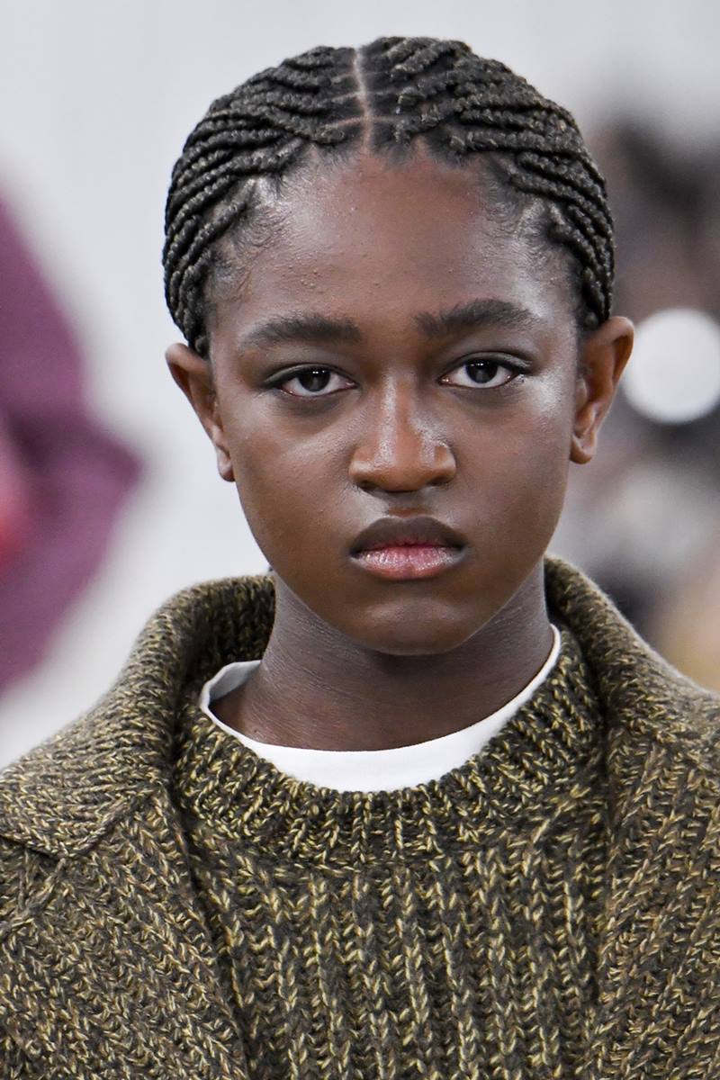 adolescente Zaya Wade, uma mulher trans e negra, de cabelos trançados pretos, no desfile da Miu Miu, na Semana de Moda de Paris. Ela usa um vestido e um casaco, ambos na cor verde, e segura uma bolsa de couro marrom. - Metrópoles