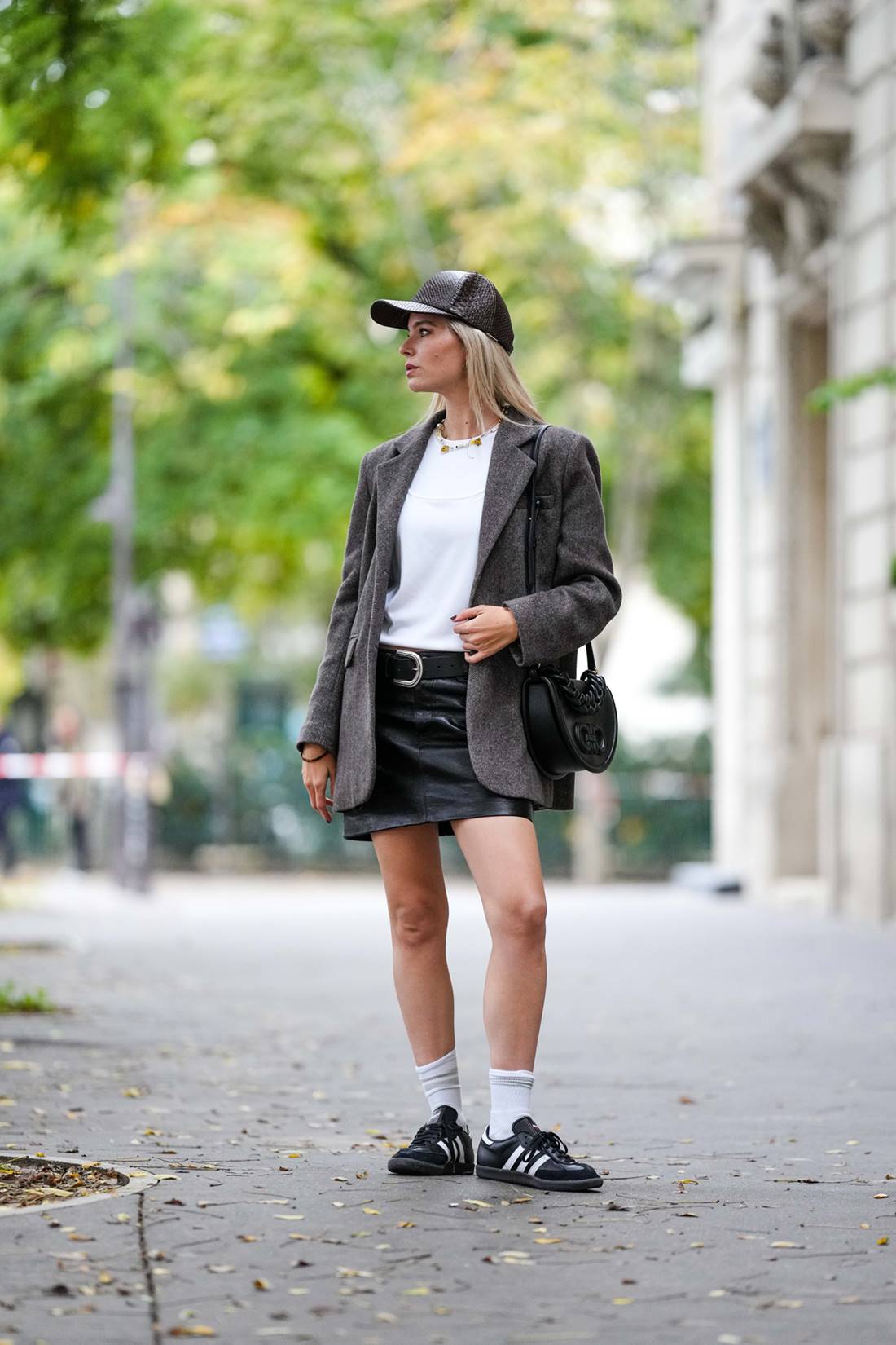 Mulher branca, jovem e loira, posa para foto na rua. Ela usa uma camiseta branca; sai e boné, ambos pretos de couro; blazer cinza; tênis Adidas, modelo Samba, na cor preta com listras brancas. - Metrópoles