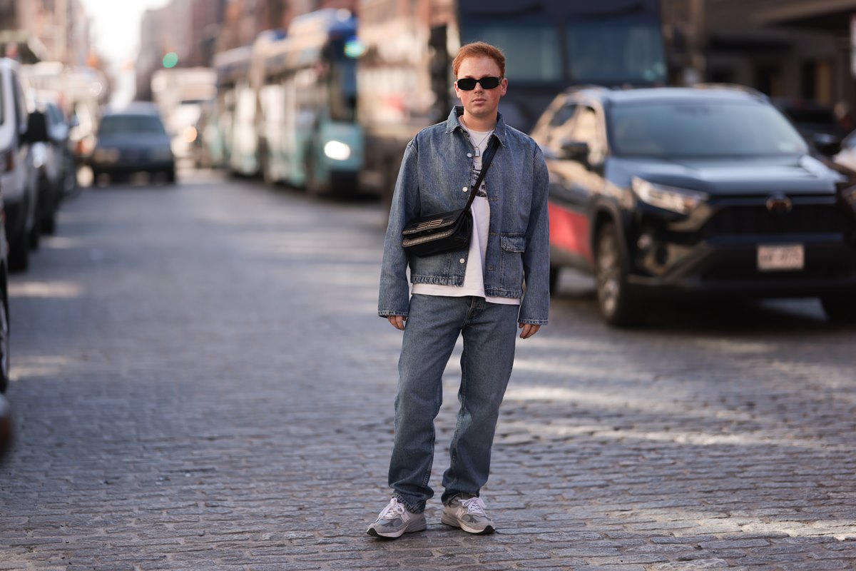 Homem branco e jovem, de cabelo curto ruivo, posa para foto no meio da rua. Ele usa uma camiseta cinza e um conjunto de jaqueta e calça, ambos jeans, em um tom também de cinza. Calça tênis de corrida da New Balence e segura uma bolsa de couro preta. - Metrópoles