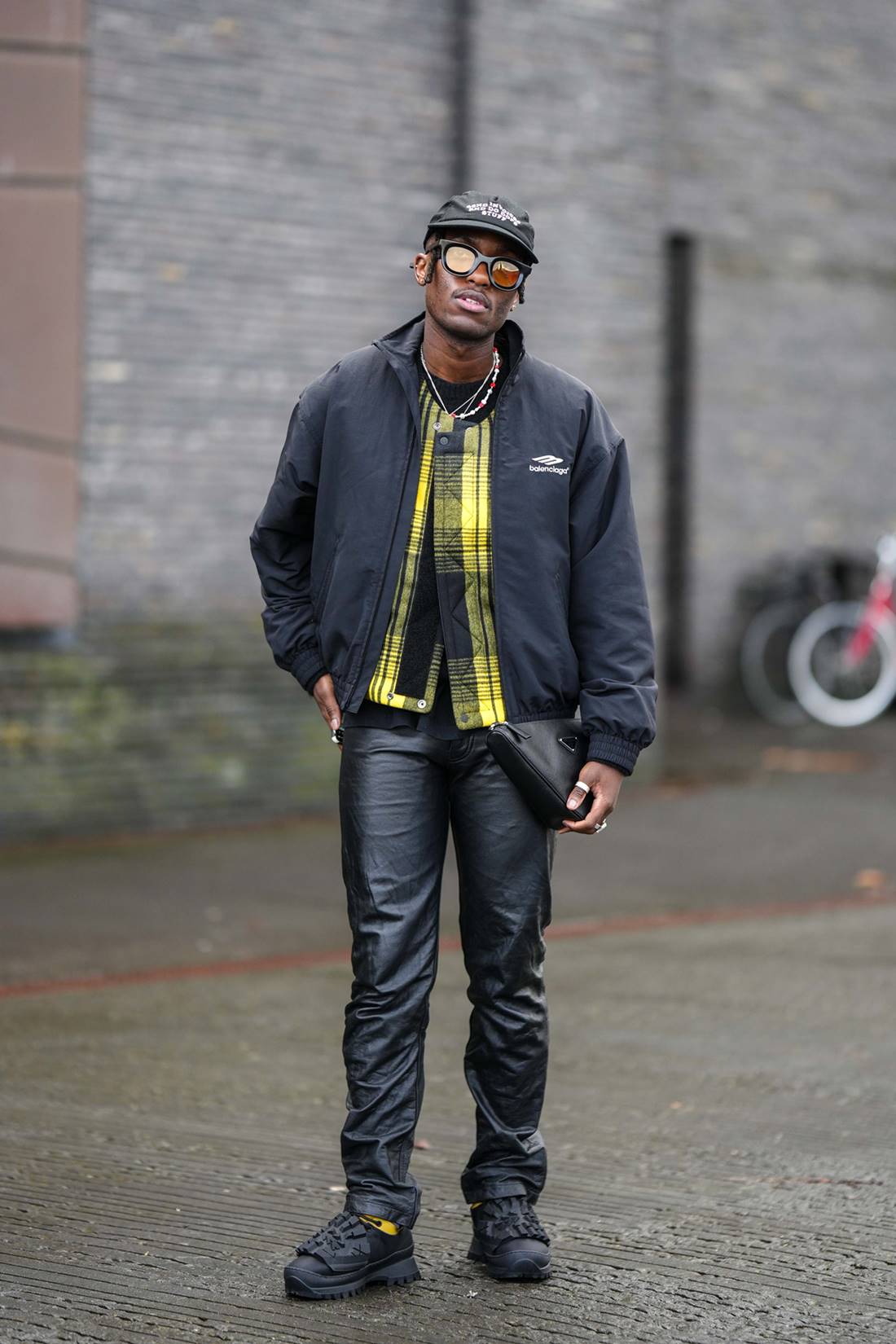 Homem jovem e negro, de cabelo trançado curto, posa para foto. Ele usa uma camisa de botão de xadrez amarelo; e um casaco e uma calça, ambos pretos, além de um tênis preto grande parecido com o de corrida. - Metrópoles