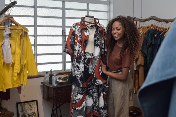 Mulher preta posa para foto em frente a uma arara de roupas. Ela está ao lado de um manequim - Metrópoles