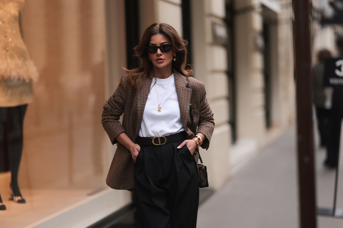 Mulher branca e jovem, de cabelo ondulado castanho longo, posa para foto na rua durante a Seman de Moda de Paris. Ela veste um camiseta branca, uma calça preta, um blazer marrom xadrez e um óculos escuro. - Metrópoles