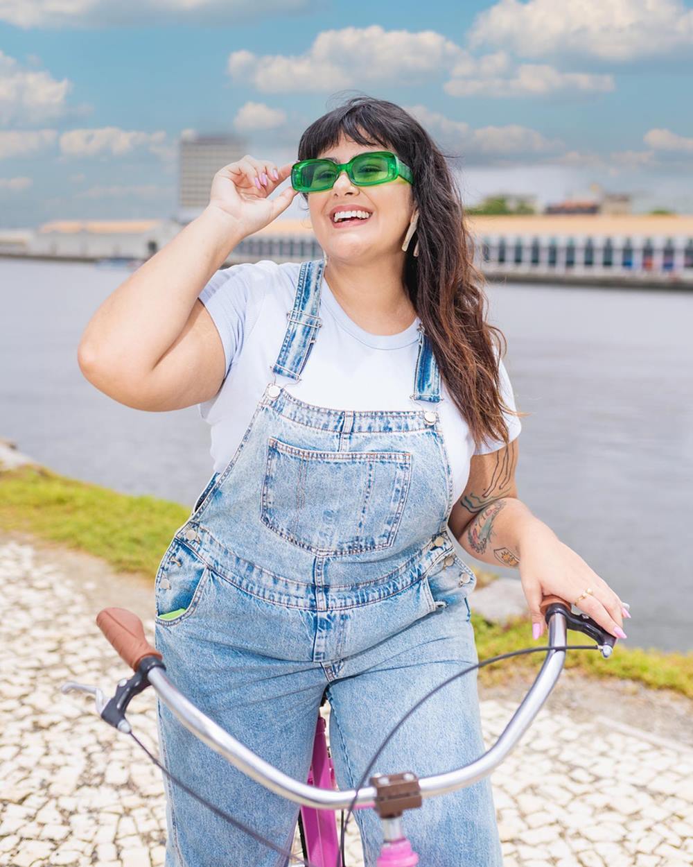 Mulher jovem e branca, com cabelo longo liso, posa para foto em uma bicicleta, no Rio de Janeiro. Usa uma camiseta branca, um macacão jeans e um óculos verde. - Metrópoles