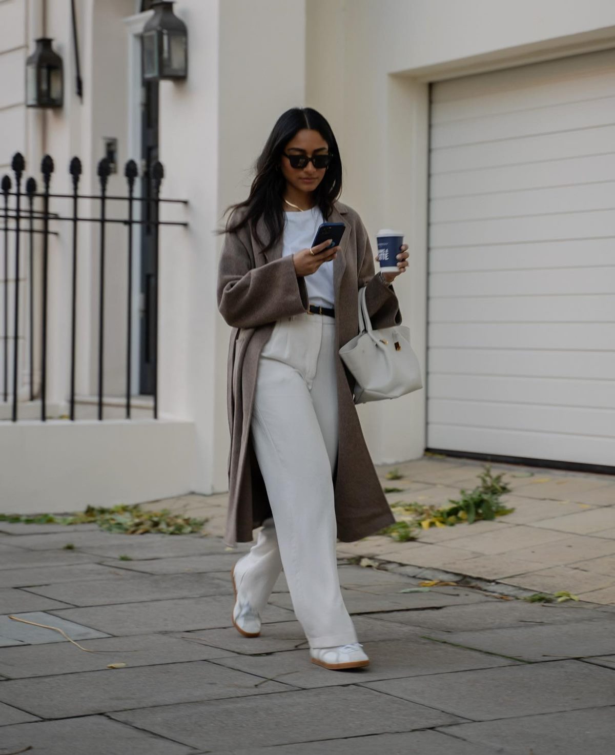 Mulher jovem e negra, com cabelo preto liso, posa para foto andando nas ruas de Londres. Usa uma camiseta branca, uma calça branca, tênis Samba da marca Adidas e um casaco bege de tricô. - Metrópoles