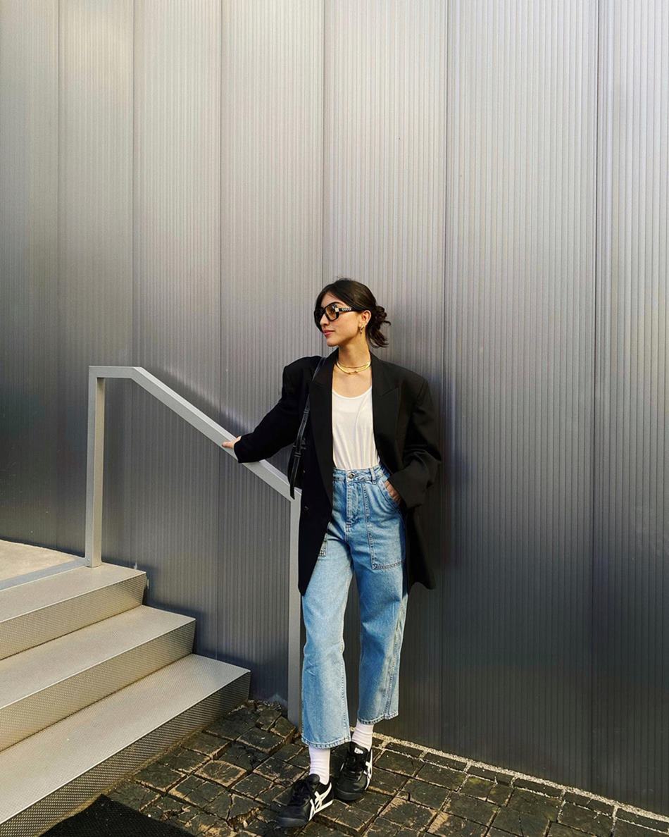 Mulher branca e jovem, de cabelo preto e liso, posa para foto em uma escada. Ela usa uma camiseta branca, uma calça jeans, um blazer preto, óculos escuros e um tênis preto. - Metrópoles