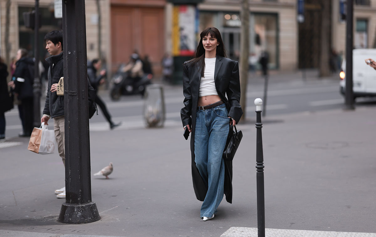 Mulher jovem e branca, de cabelo liso preto, anda pelas ruas durante a Semana de Moda de Paris. Ela usa uma camiseta branca cropped, calça jeans ampla, sandália branca de salto e um casaco longo, preto e de couro. - Metrópoles