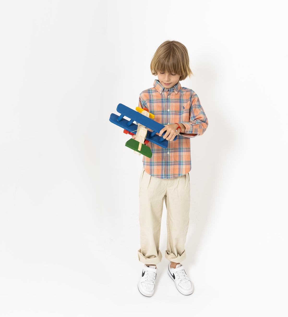 Menino criança, de no máximo 8 anos, branco e com cabelo liso loiro. Ele posa para campanha do brechó Prettynew e usa uma camisa de botão xadrez, uma calça bege e um tênis branco. - Metrópoles