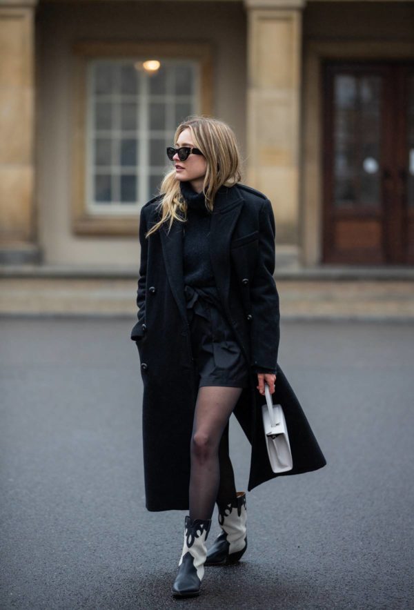 Mulher jovem e loira, de cabelo longo liso, andando pelas ruas de Berlim, na Alemanha. Ela usa óculos escuros, um tricô preto de gola alta, shors preto, bota de cowboy preta e branca e um casaco trench coat preto. - Metrópoles