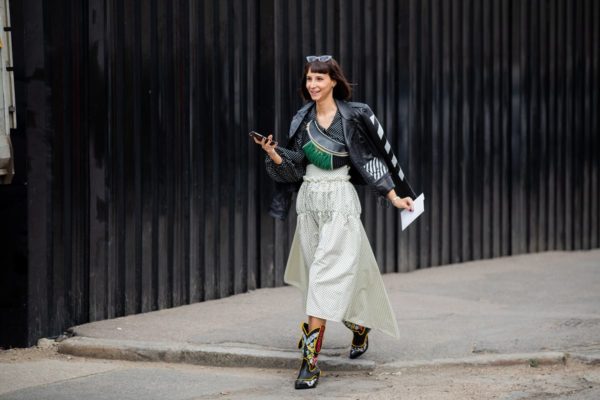 Mulher branca e jovem, de cabelo liso preto, anda pela durante a semana de moda. Ela usa blusa preta, saia longa branca, jaqueta de couro e bota preta de cowboy com detalhes coloridos. - Metrópoles