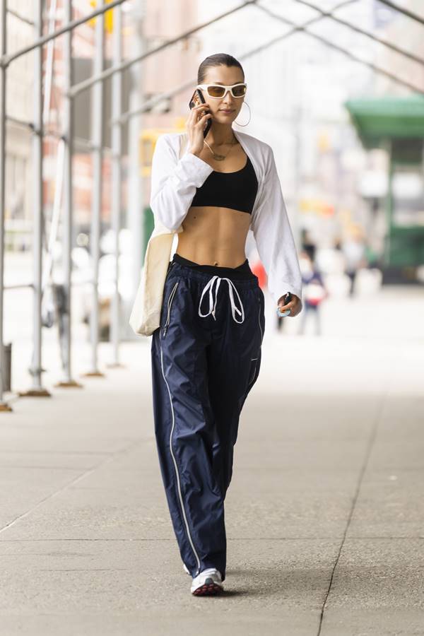 Bella Hadid falando ao celular. Ela usa top preto, camisa branca e calça paraquedas azul esportiva - Metrópoles