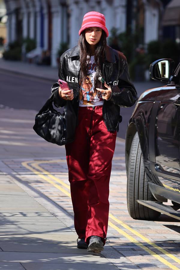 Na rua, Dua Lipa usa chapéu rosa, calça paraquedas vermelha, camiseta branca e jaqueta preta - Metrópoles