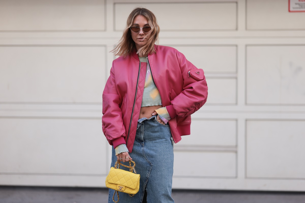 Mulher branca e jovem, de cabelo liso loiro, posa para foto na rua. Ela usa um top cropped verde; uma saia jeans; jaqueta bomber rosa de couro e uma bolsa amarela de couro, amarela, da Chanel. - Metrópoles