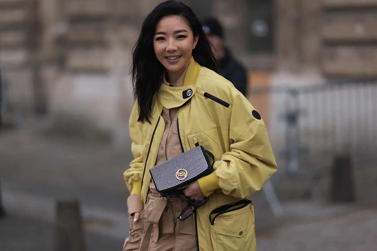 Mulher asiática, branca e jovem, de cabelo preto liso, posa para foto durante a semana de moda. Ela usa um macacão bege, uma jaqueta bomber amarela com zíperes preto, uma bolsa preta com strass. - Metrópoles