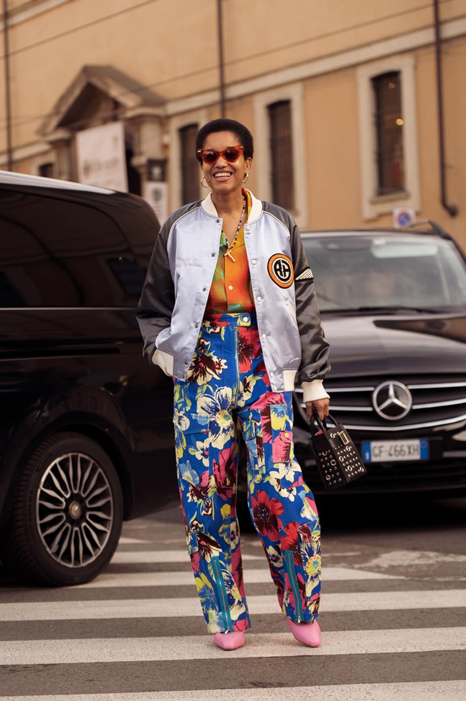 Mulher negra e de meia idade, com cabelo crespo curto, posa para foto na Semana de Moda de Milão. Ela usa uma camisa de botão laranja; uma calça azul com flores estampadas; uma jaquta bomber prateada e sapatos rosas de couro. - Metrópoles