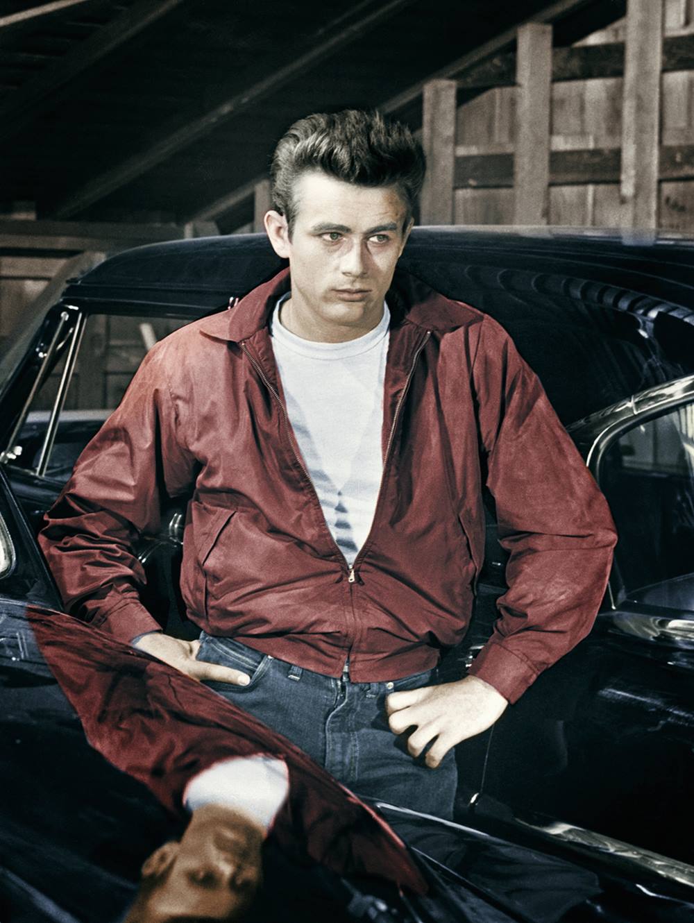 O ator James Dean, um homem branco e jovem, de cabelo castanho liso em topete, em cena de filme. Está encostado em um carro e usa camiseta branca, calça jeans e uma jaqueta bomber vermelh de couro. - Metrópoles