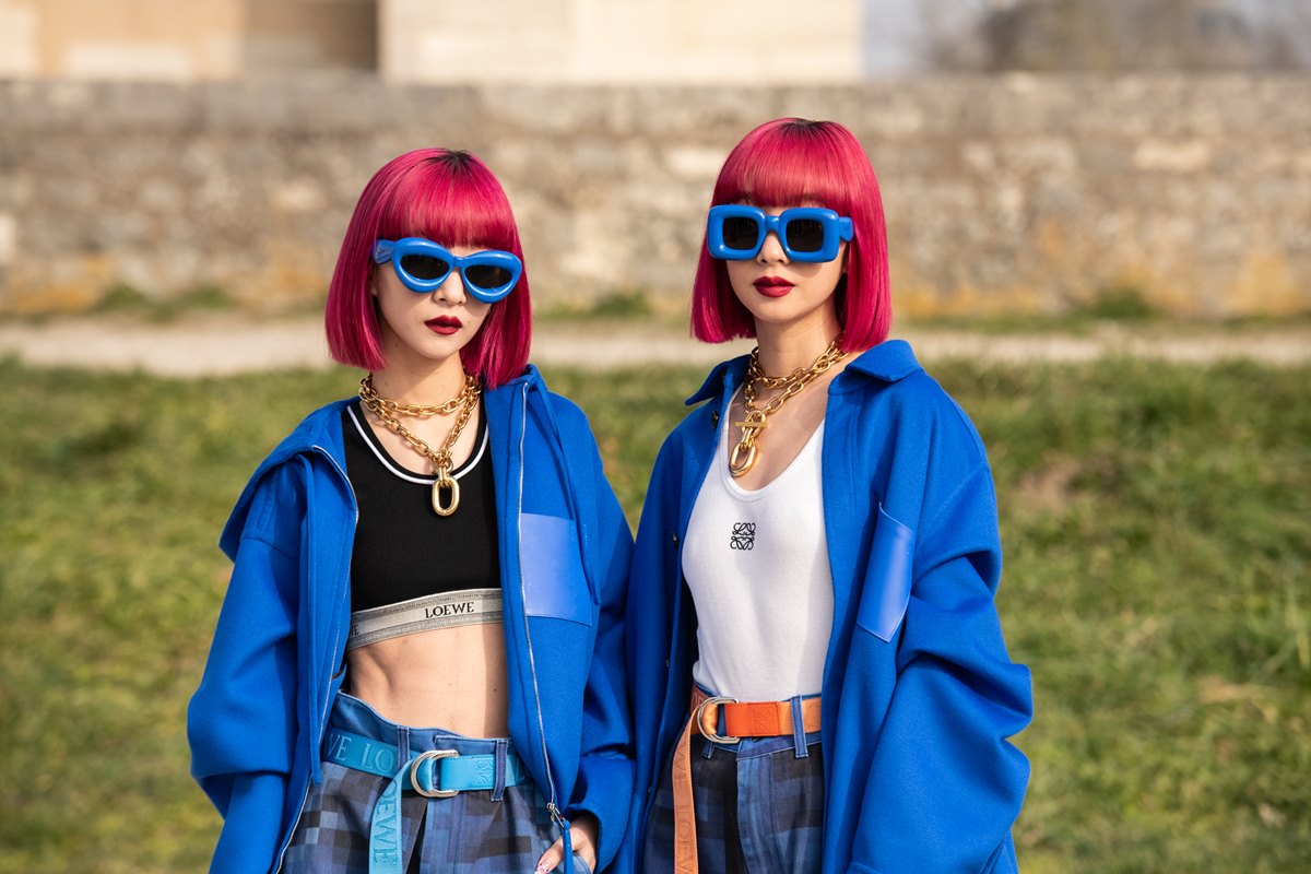 Duas irmãs gêmeas asiáticas, brancas de cabelo liso tingido de rosa, posam para foto no desfile da Loewe na Semana de Moda de Paris. Elas usam calças jeans folgadas e óculos de sol e casaco, ambos azul. Uma usa um top preto curto e a outra uma regata branca. - Metrópoles