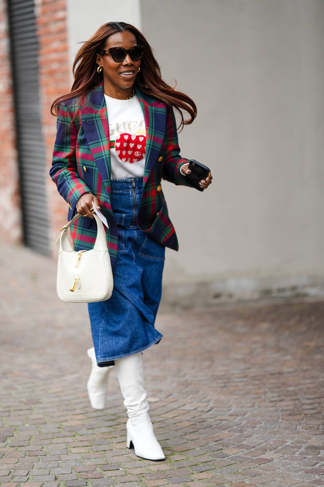 Mulher negra e jovem, de cabelo castanho liso, andando pela rua durante a Semana de Moda de Milão. Ela usa uma camiseta da Gucci, uma saia jeans, um blazer xadrez e botas de salto alto brancas. - Metrópoles