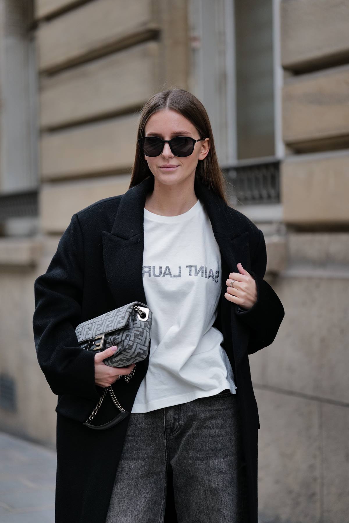 Mulher branca e jovem, de cabelo castanho liso e longo, posa para foto na rua durante a Semana de Moda de Paris. Ela usa uma camiseta branca, com a palavra Saint Laurent bordada, uma calça jeans preta, casaco preto longo e óculos escuros. - Metrópoles