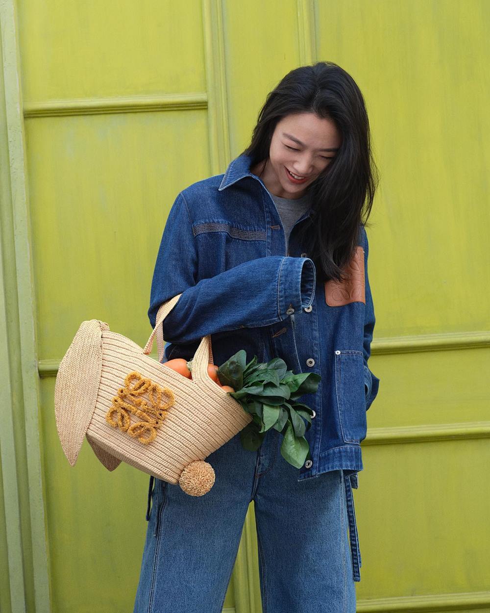 Mulher branca, asiática e jovem, de cabelo liso preto, posa para foto de campanha da grife Loewe. Ela usa jaqueta e calça, ambas no tecido jeans, e uma bolsa de palha bege. - Metrópoles