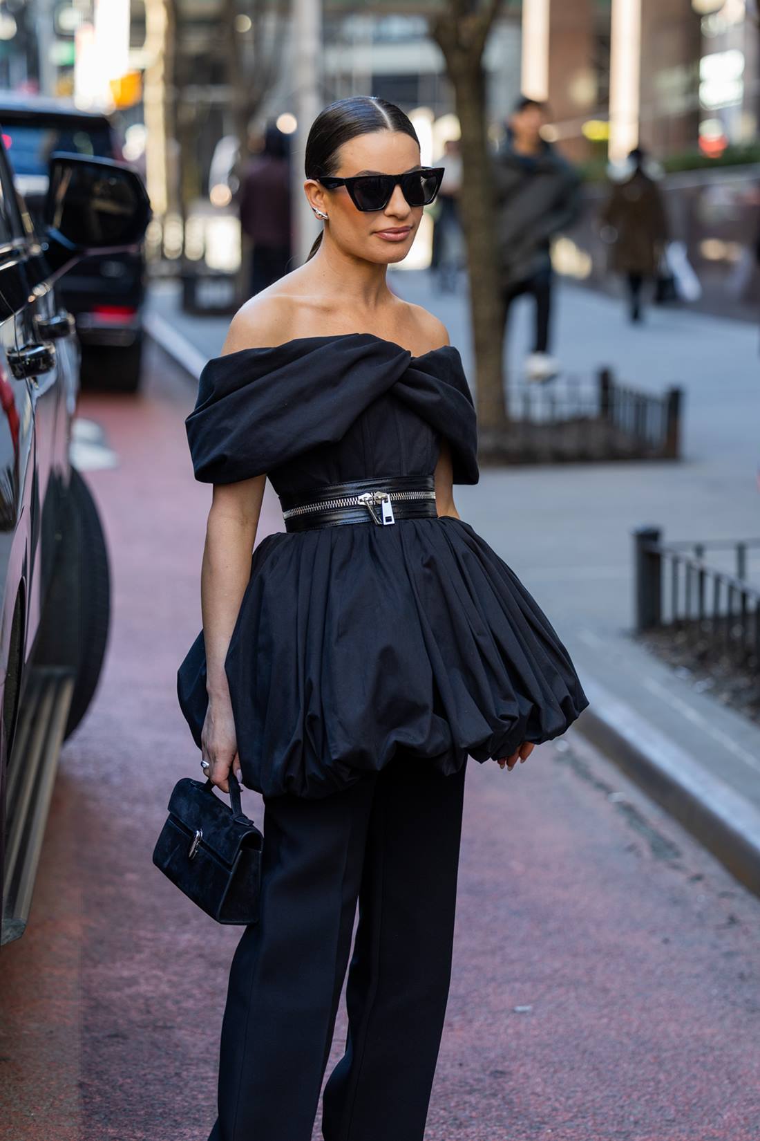 A atriz Lea Michele, uma mulher branca e jovem, com cabelo liso preto, posa para foto na rua. Ela usa um vestido curto preto e uma calça reta, também preta, além de óculos escuros. - Metrópoles