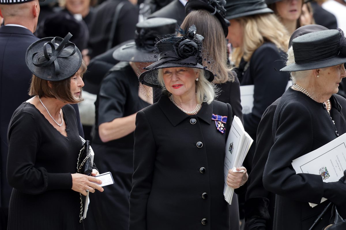 Foto colorida de mulher idosa e branca ao redor de várias pessoas vestidas de preto - Metrópoles