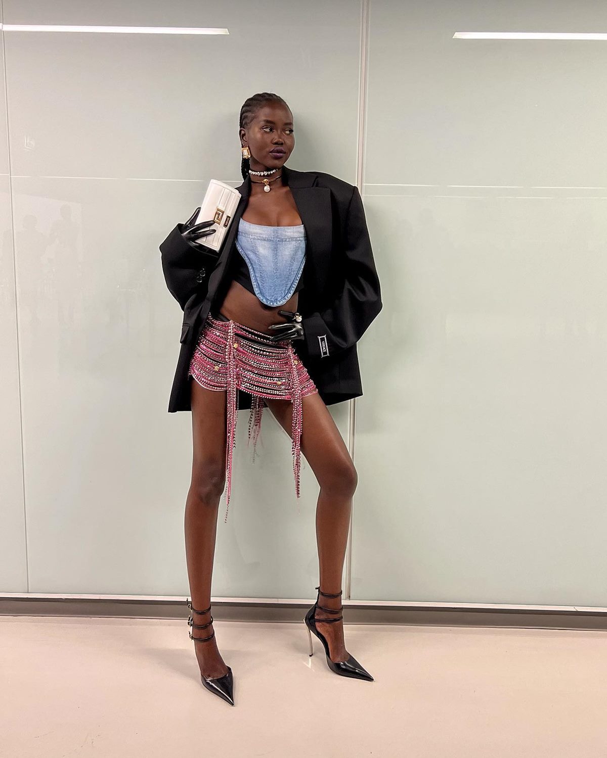 Um modelo mulher, jovem e negra, com cabelo preto trançado, posa para foto em fundo branco. Ela veste um top curto jeans, saia rosa de brilho, blazer preto e sandália preta de salto. - Metrópoles