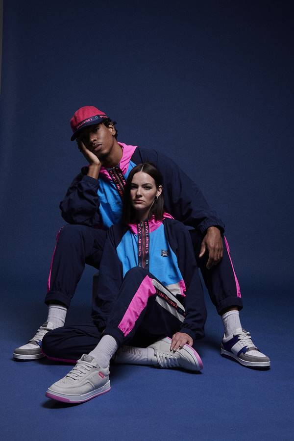 Dois modelos posando para foto de moda. O fundo é azul e eles estão usando roupas esportivas - Metrópoles