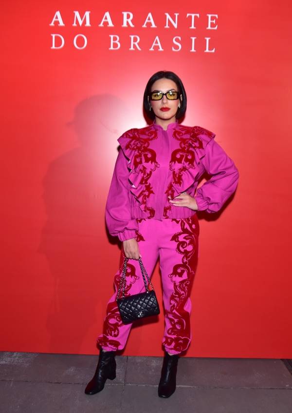 Mulher com roupa rosa posando para foto. Ela está segurando a cintura com uma das mãos e em frente a um fundo vermelho - Metrópoles