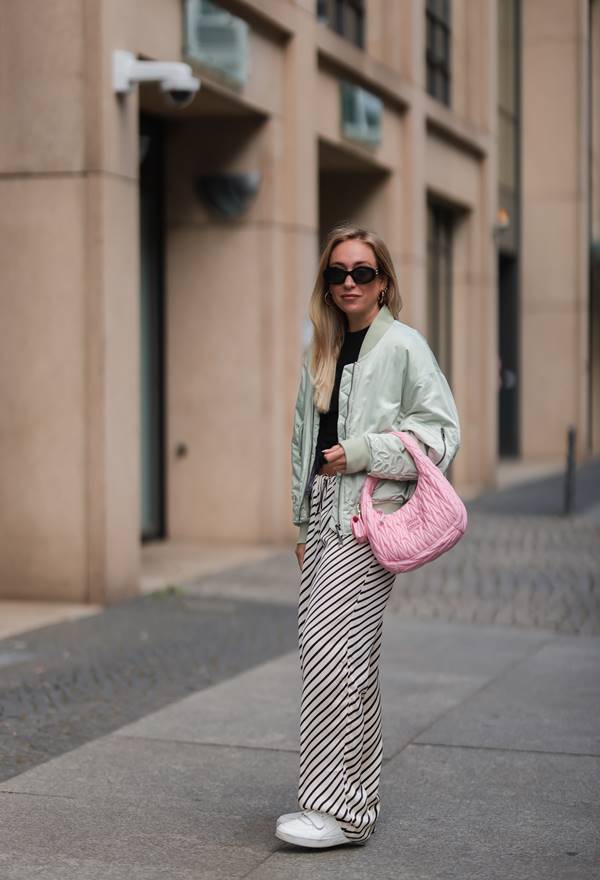 Mulher na rua posando para foto. Ela está usando óculos de sol, jaqueta grande e com a bolça apoiada no braço - Metrópoles