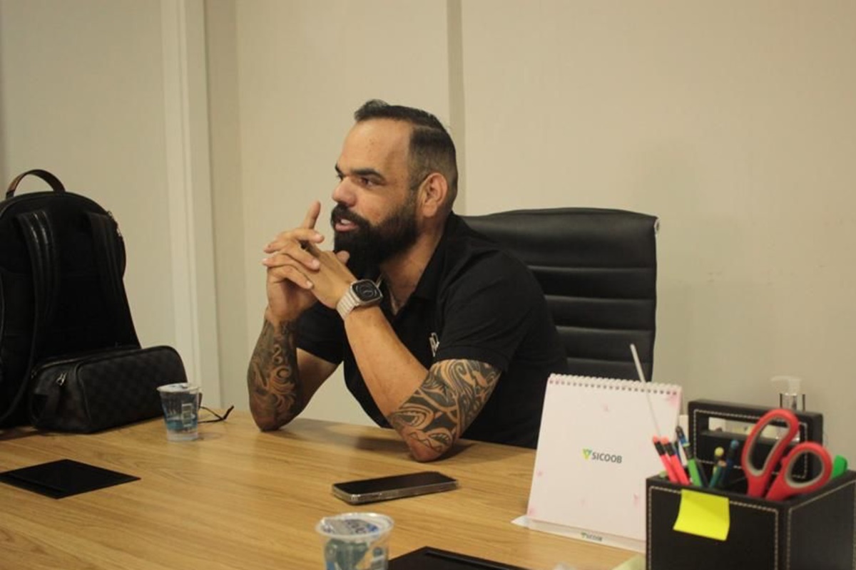 Foto colorida de homem pardo, de barba, sentado em frente a uma mesa com itens de papelaria e mochila - Metrópoles