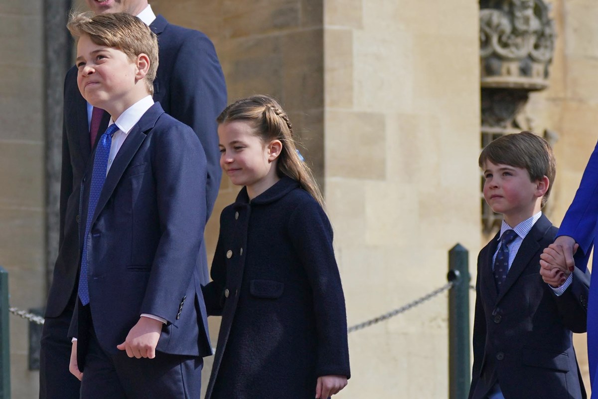 Foto colorida de menino loiro, branco e de terno à frente de uma menina loira, branca e com casaco preto. Atrás está um menino branco, loiro e com blazer e gravata - Metrópoles