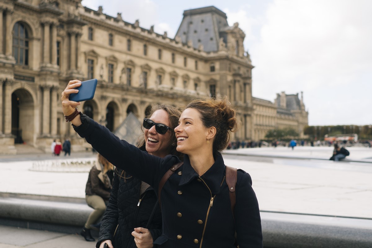 Foto colorida de duas mulheres brancas tirando uma selfie em frente ao Museu do Louvre - Metrópoles