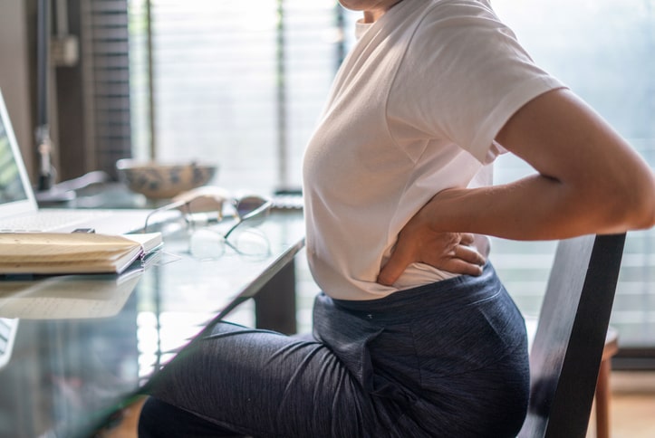 Imagem colorida mostra mulher sentada em escritório, com a mão nas costas, se alongando - Metrópoles