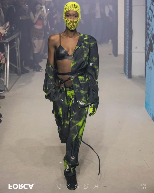 Na imagem com cor, uma mulher negra com uma balaclava verde neon no rosto usando um conjunto de camisa e calça verde e preto estampado desfilando - Metrópoles