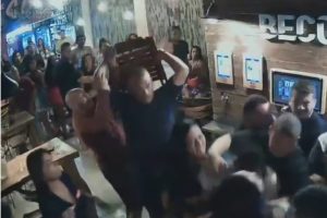 várias pessoas em um bar, homem com as mãos para trás segura cadeira