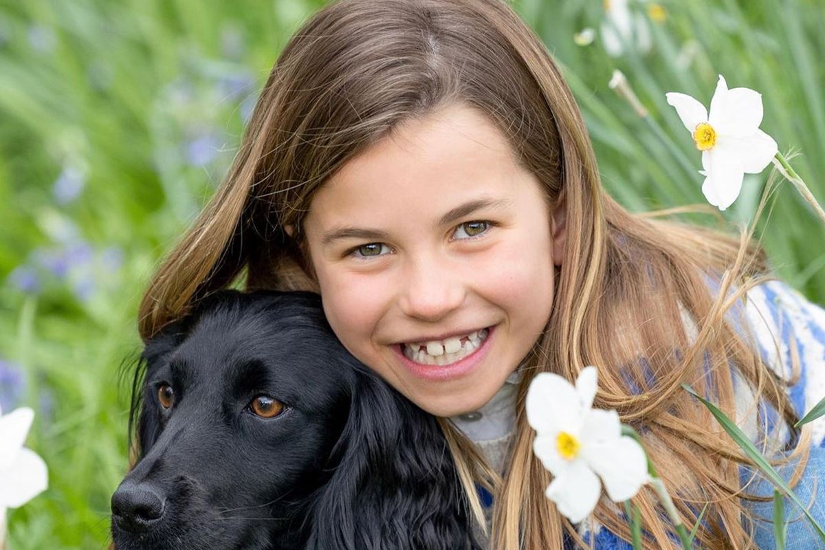 Foto colorida de menina branca e loira abraçada a um cachorro de pelo preto. Ela e o cão estão em um jardim - Metrópoles