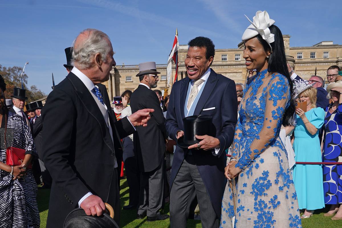 Foto colorida de um homem idoso e branco ao lado de um homem e uma mulher negros. Eles conversam e estão ao lado de um grupo de pessoas - Metrópoles