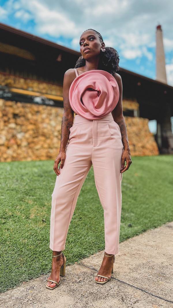 Com grama ao fundo, mulher negra posa usando look rosa composto por calça e top com flor em 3D - Metrópoles