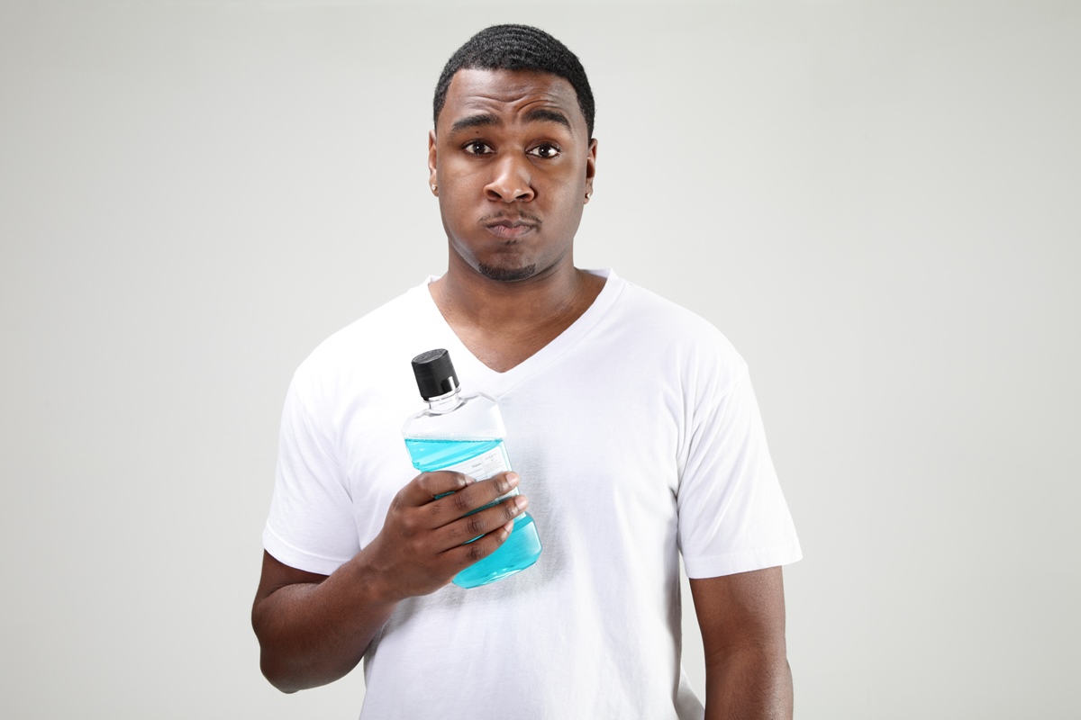 Foto colorida de homem negro segurando um frasco de enxaguante bucal e ele está com o produto na boca - Metrópoles