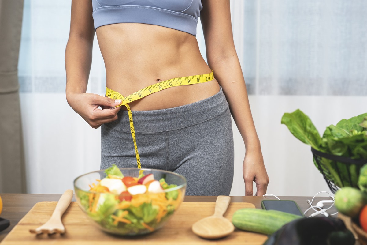 Foto colorida de mulher com fita métrica amarela na cintura. Perto dela tem uma colher de pau e uma bacia com vegetais - Metrópoles