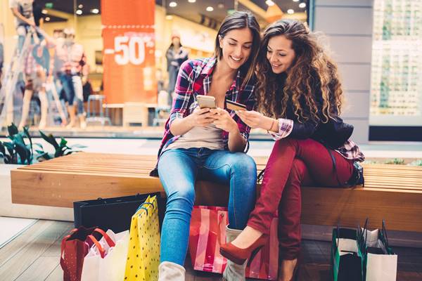 Duas jovens mulheres, sentadas em um banco no shopping, fazendo compras on-line - Metrópoles