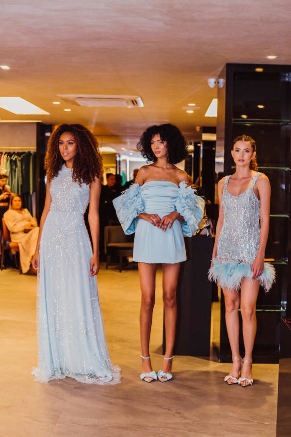 Na imagem com cor, três mulheres, dua delas negras e uma branca, posando em uma loja com vestidos em azul claro - Metrópoles