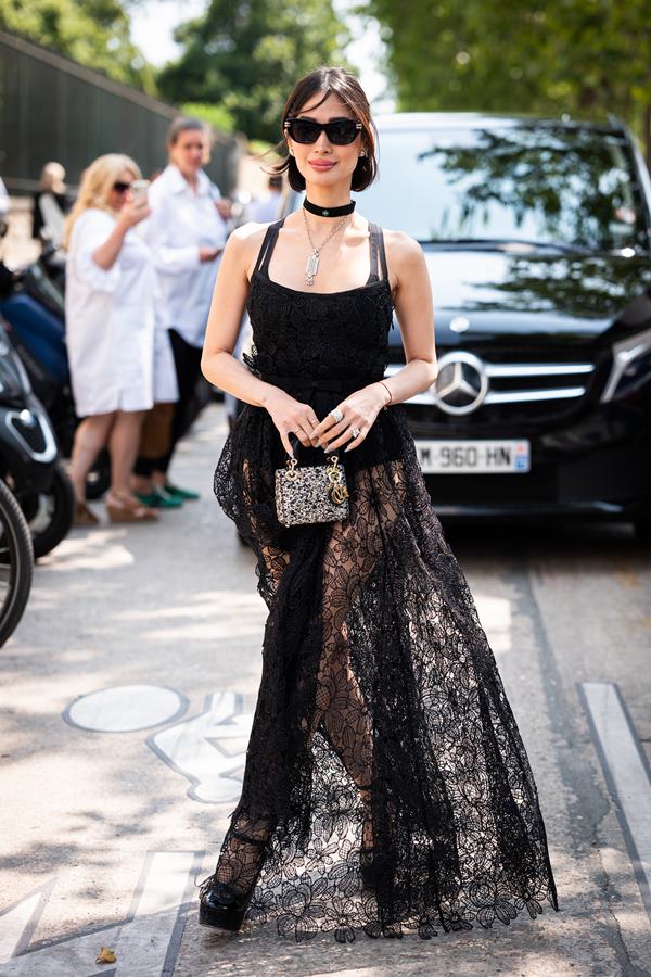 Mulher andando na rua com vestido preto. Ela está segurando uma bolsa pequena com as duas mãos - Metrópoles